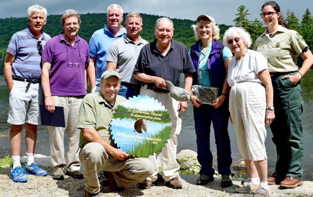 Now-retired Androscoggin District Ranger Katie Stuart of Shelburne, 3rd from right, holds the plaque honoring her collaboration with the Randolph Community Forest presented to her on Aug. 6 by Randolph town moderator David Willcox, 4th from right, at the Pond of Safety. Also honoring her: Forest commissioner Walter Graff, left, Chuck Henderson representing U.S. Sen. Jeanne Shaheen, state Sen. Jeff Woodburn, WMNF Supervisor Tom Wagner, Rep. Paula Bradley, prime sponsor of legislation creating the Commission & Forest, and today’s Androscoggin District Ranger Jennifer Barnhart. Randolph Forest Commission chairman John Scarinza, front row, holds the saw blade he presented to Stuart. (Edith Tucker photo) 